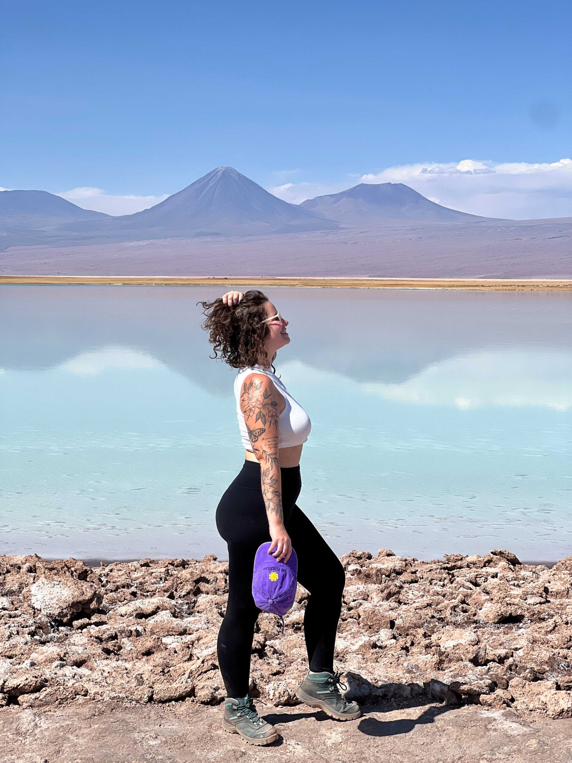 Mulher na margem de uma lagoa de agua turquesa com vulcoes ao fundo no deserto do Atacama, Chile