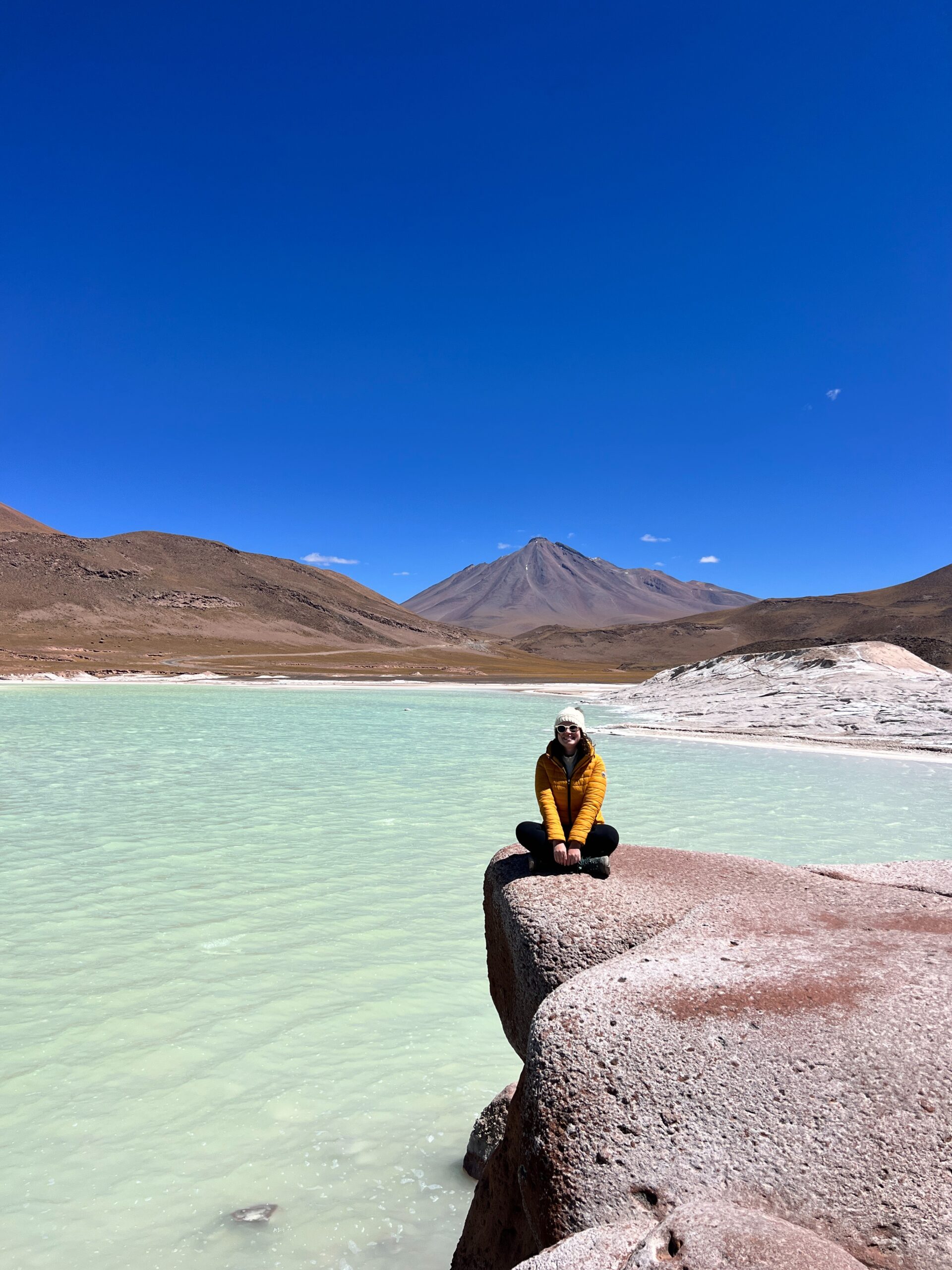 Mulher sentada em uma rocha avermelhada diante da lagoa de agua turquesa de Piedras Rojas, no deserto do Atacama, Chile