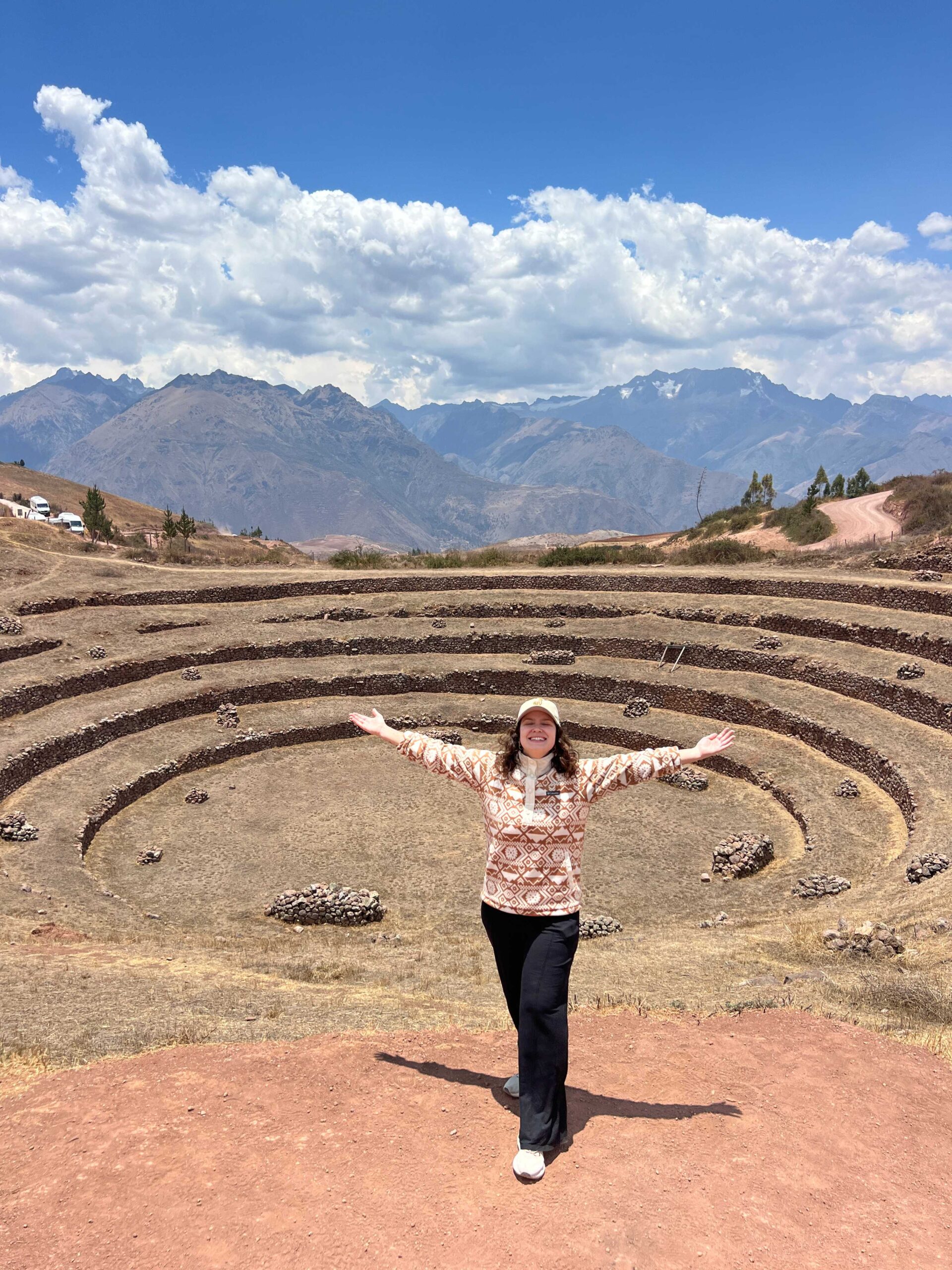 Mulher de braços abertos diante dos terraços circulares incas de Moray, no Vale Sagrado, com os Andes ao fundo