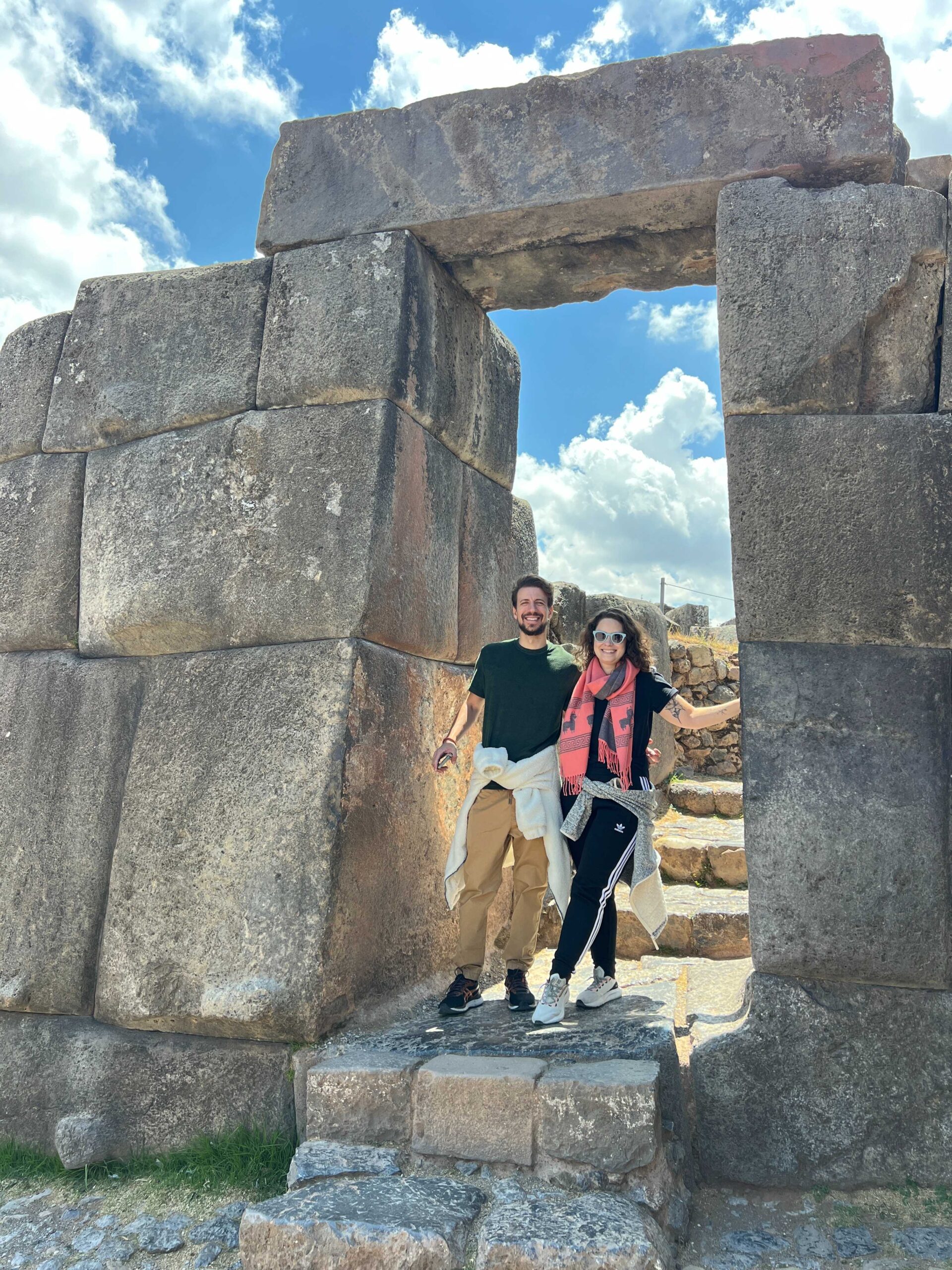 Casal em pé sob o portal de pedras gigantes da fortaleza inca de Sacsayhuamán, em Cusco, no Peru