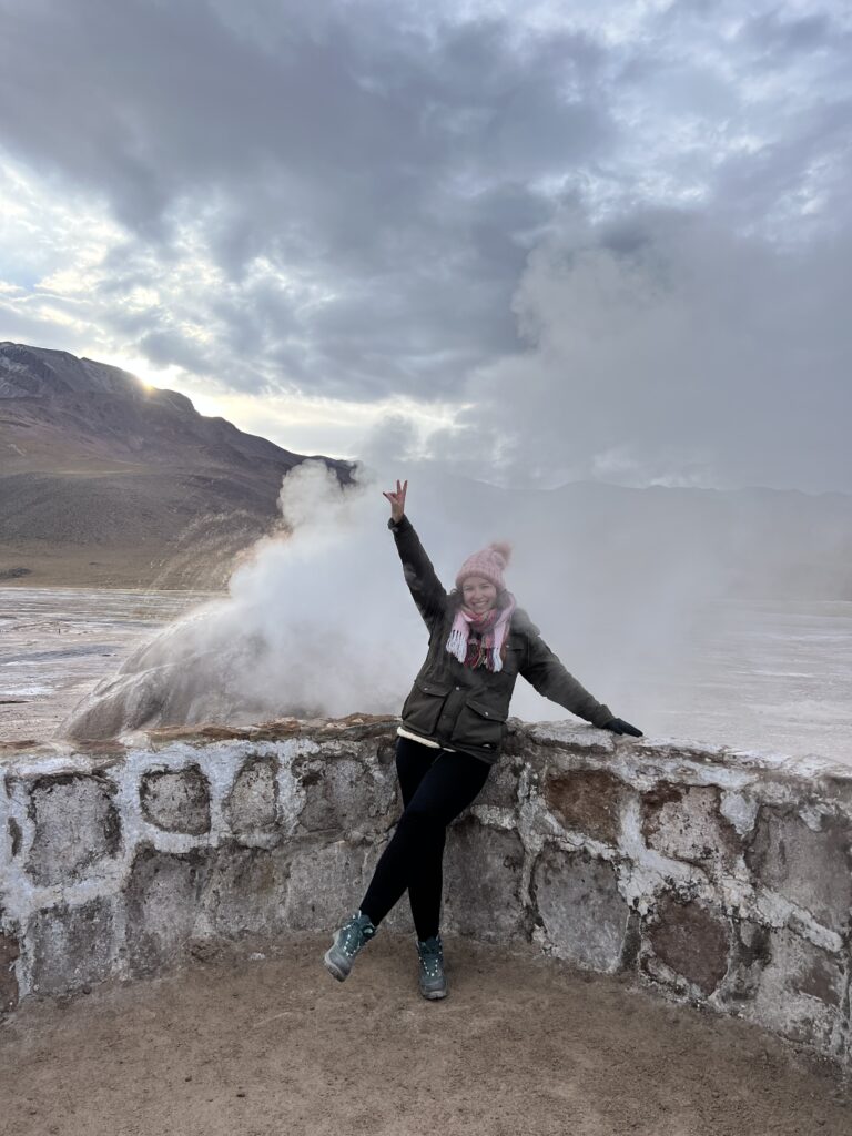 Mulher de braços abertos ao lado de uma fumarola dos Geiseres del Tatio, Atacama