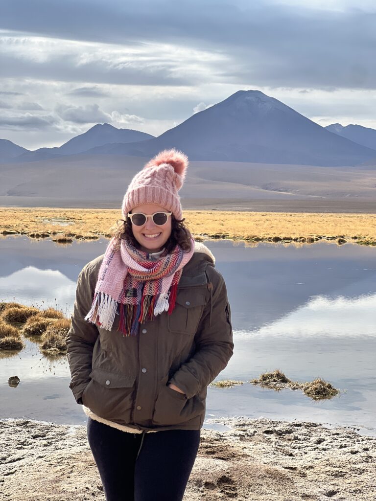 Mulher de gorro rosa diante de laguna altiplânica com vulcão ao fundo no Atacama