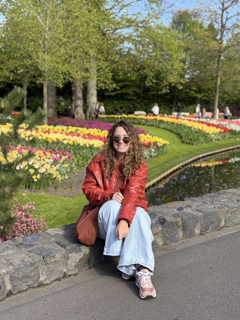 Jéssica sentada em um muro de pedra no Keukenhof, com canteiros de tulipas coloridas e o lago do jardim ao fundo